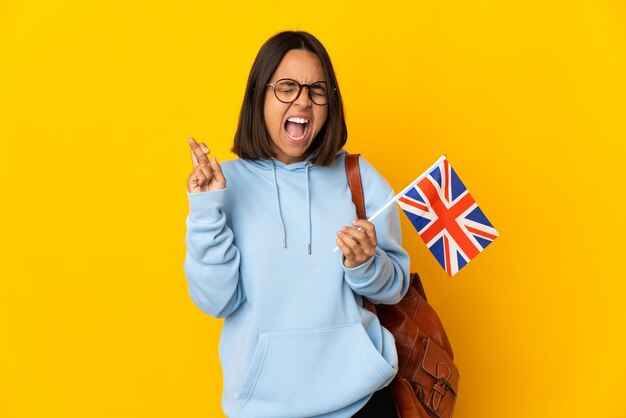 Jeune femme latine tenant un drapeau du Royaume-Uni isolé sur un mur jaune avec des doigts traversant