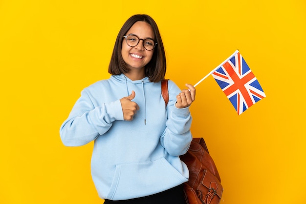 Jeune femme latine tenant un drapeau du Royaume-Uni isolé sur un mur jaune donnant un geste de pouce en l'air