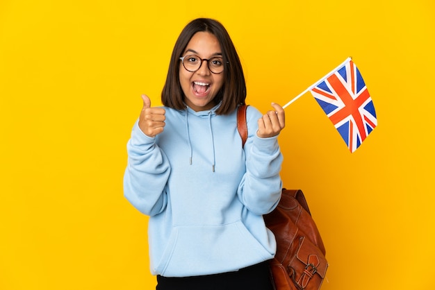 Jeune femme latine tenant un drapeau du Royaume-Uni isolé sur un mur jaune montrant le signe ok et le geste du pouce vers le haut