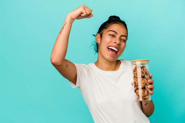 Jeune femme latine tenant un pot de biscuits isolé sur fond bleu levant le poing après une victoire, concept gagnant.