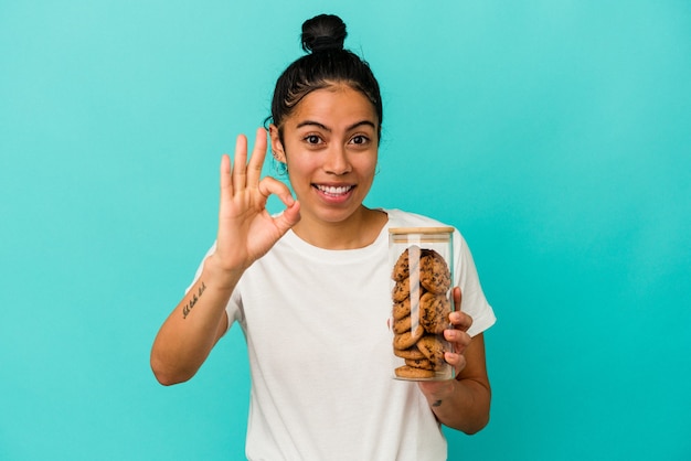 Jeune femme latine tenant un pot de cookies isolé sur fond bleu joyeux et confiant montrant un geste ok.