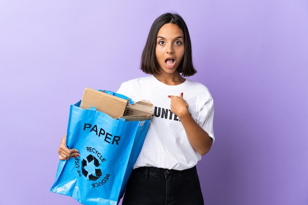 Jeune femme latine tenant un sac de recyclage plein de papier à recycler isolé sur violet avec expression faciale surprise
