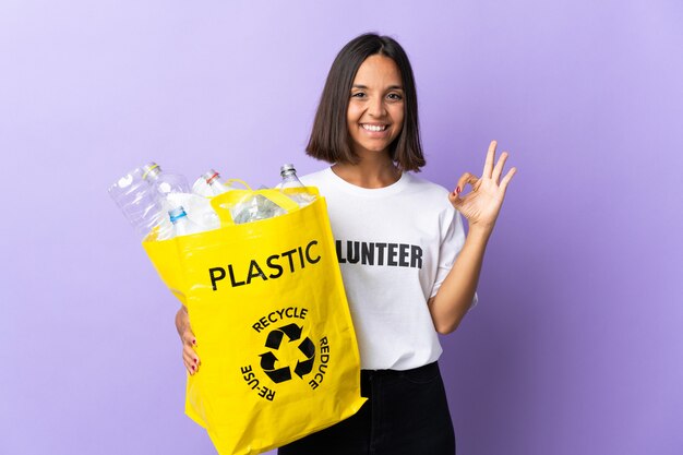 Jeune femme latine tenant un sac de recyclage plein de papier à recycler isolé sur violet montrant signe ok avec les doigts