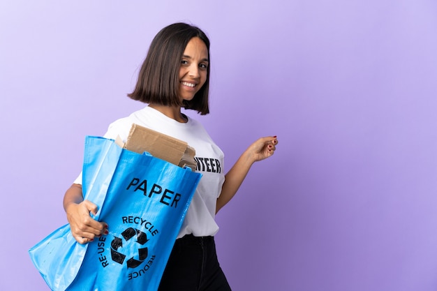 Jeune femme latine tenant un sac de recyclage plein de papier à recycler isolé sur violet pointant vers l'arrière