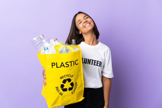 Jeune femme latine tenant un sac de recyclage plein de papier à recycler isolé sur violet en riant