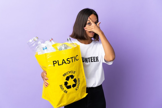 Jeune femme latine tenant un sac de recyclage plein de papier à recycler isolé sur les yeux cône violet par les mains et souriant