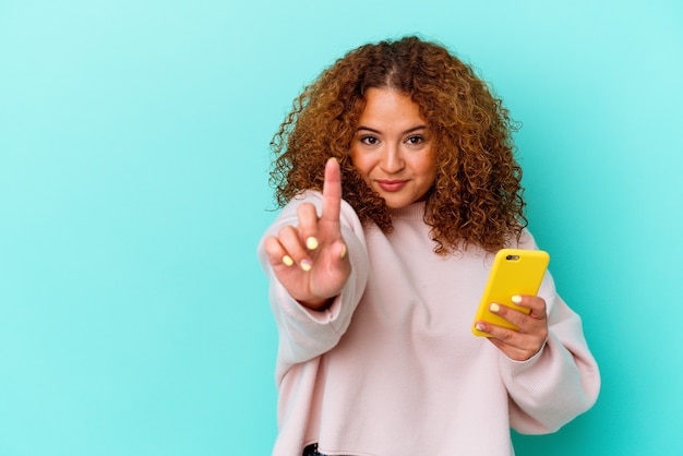 Jeune femme latine tenant un téléphone mobile isolé sur fond bleu montrant le numéro un avec le doigt.