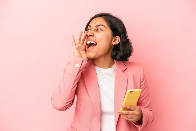 Jeune femme latine tenant un téléphone portable isolé sur fond rose criant et tenant la paume près de la bouche ouverte.