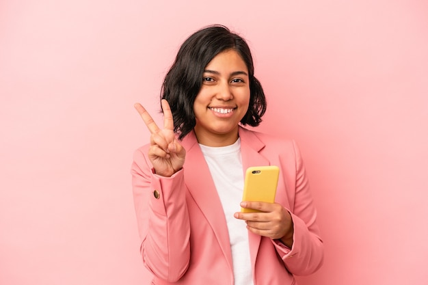 Jeune femme latine tenant un téléphone portable isolé sur fond rose montrant le numéro deux avec les doigts.