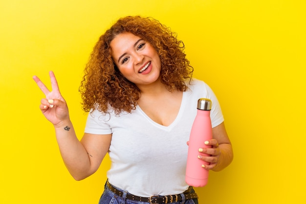 Jeune femme latine tenant un thermos isolé sur fond jaune joyeux et insouciant montrant un symbole de paix avec les doigts.