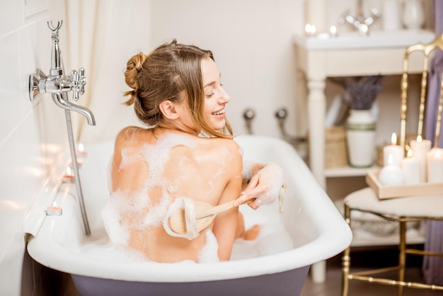 Jeune femme lavant avec une brosse dans la belle baignoire vintage pleine de mousse dans la salle de bain décorée de bougies