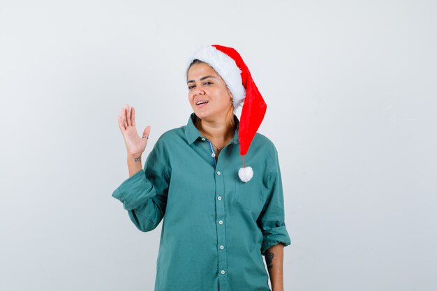 Jeune femme levant la main dans un chapeau de Noël, une chemise et l'air gai. vue de face.