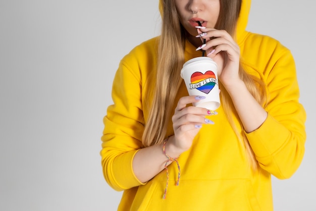 Jeune femme LGBTQ portant une veste à capuche jaune, boire du café dans une tasse avec coeur arc-en-ciel.