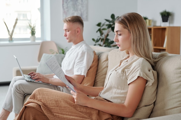 Jeune femme lisant un contrat commercial ou un accord éducatif contre son mari en train de taper sur un ordinateur portable alors qu'elles étaient toutes les deux assises sur un canapé à la maison