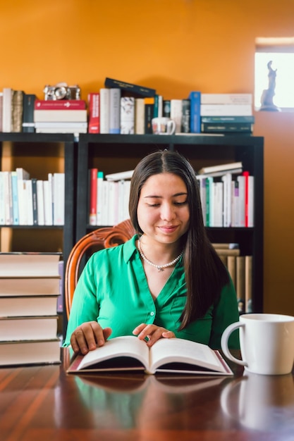 Jeune femme lisant un livre dans sa bibliothèque personnelle avec une étagère derrière elle, avec une tasse à café.