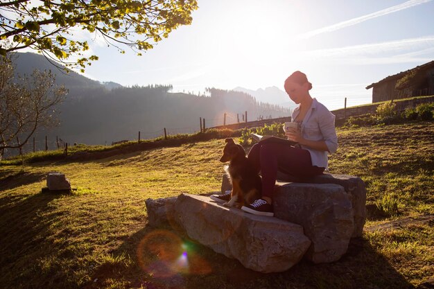 Jeune femme lisant un livre à la lumière de l'aube avec une tasse de café à la main et la compagnie de son chien bodeguero Countryside Copy space