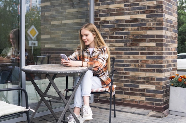 Une jeune femme lisant un message drôle au téléphone alors qu'elle est assise à une table de cafétéria en plein air