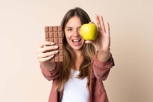 Jeune femme lituanienne sur beige prenant une tablette de chocolat dans une main et une pomme dans l'autre