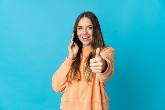 Jeune femme lituanienne isolée sur mur bleu en gardant une conversation avec le mobile tout en faisant les pouces vers le haut