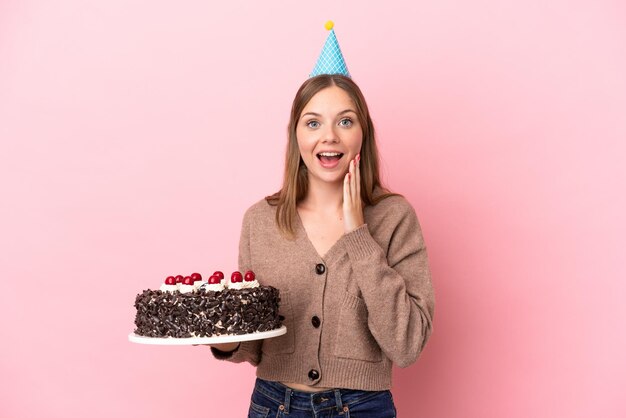 Jeune femme lituanienne tenant un gâteau d'anniversaire isolé sur fond rose avec une expression faciale surprise et choquée