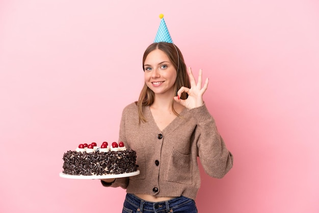 Jeune femme lituanienne tenant un gâteau d'anniversaire isolé sur fond rose montrant un signe ok avec les doigts