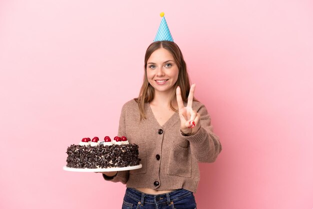 Jeune femme lituanienne tenant un gâteau d'anniversaire isolé sur fond rose souriant et montrant le signe de la victoire