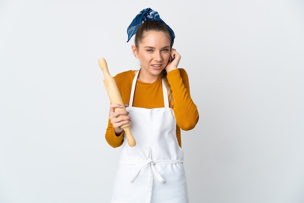 Jeune femme lituanienne tenant un rouleau à pâtisserie isolé sur un mur blanc frustré et couvrant les oreilles