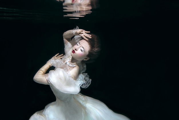 jeune femme en longue robe de luxe blanche sous l'eau sur le fond noir. Mouvement, art, hiver