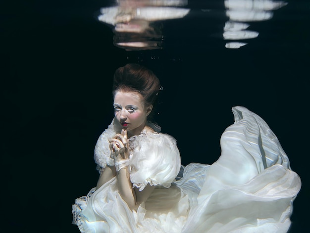 jeune femme en longue robe de luxe blanche sous l'eau sur le fond noir. Mouvement, art, hiver