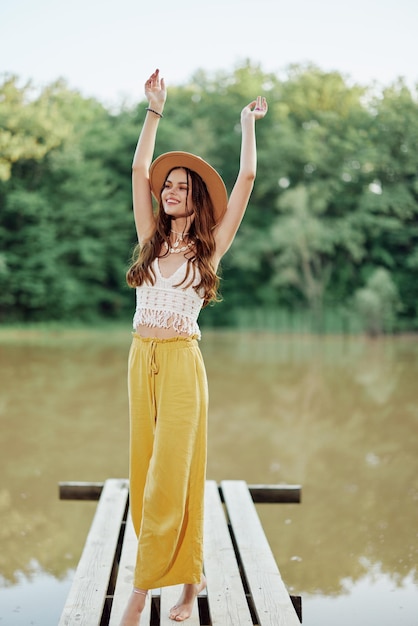 Une jeune femme en look hippie et écodress voyage dans la nature au bord du lac portant un chapeau et un pantalon jaune au coucher du soleil d'automne Photo de haute qualité