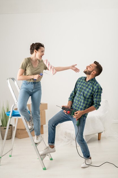 Jeune femme ludique chantant dans le rouleau à peinture tandis que son mari à l'aide de la perceuse comme guitare, ils s'amusent tout en faisant la rénovation