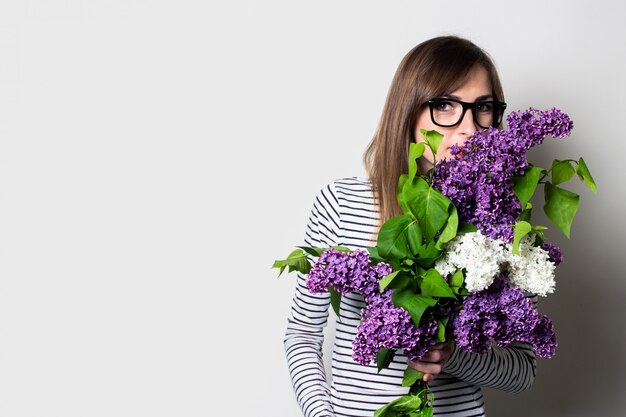 Jeune femme à lunettes aime l'arôme d'un bouquet de lilas sur un fond clair.