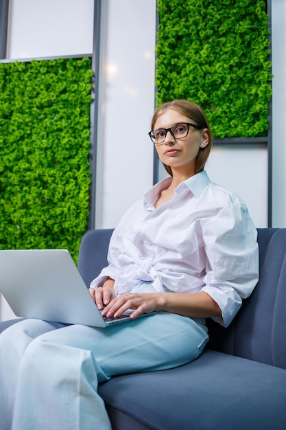 Une jeune femme à lunettes et une chemise en coton blanc est assise sur un canapé et travaille en ligne sur un ordinateur portable