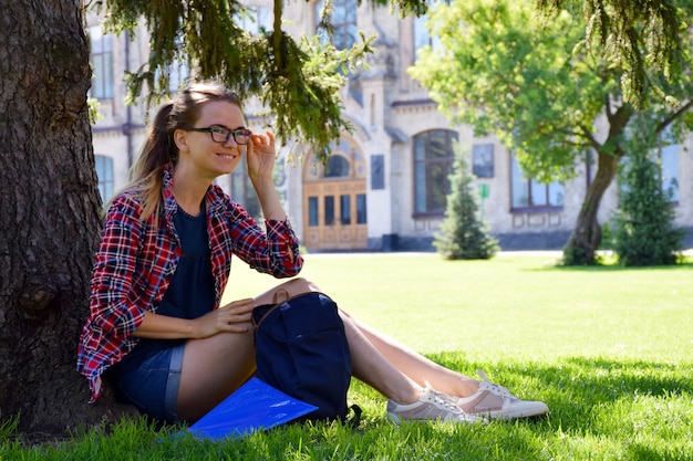 Jeune femme à lunettes est assise sur l'herbe sous l'arbre près de l'université.