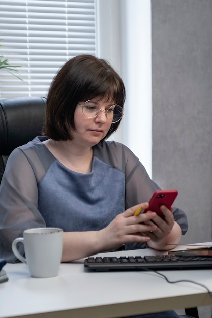 Jeune femme à lunettes est assise sur le lieu de travail et regarde le smartphone. Gestionnaire SMM travaillant. Animation des réseaux sociaux. Cadre vertical