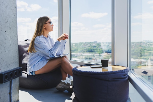 Une jeune femme à lunettes est assise sur un pouf doux près de la fenêtre et regarde la ville depuis un gratte-ciel