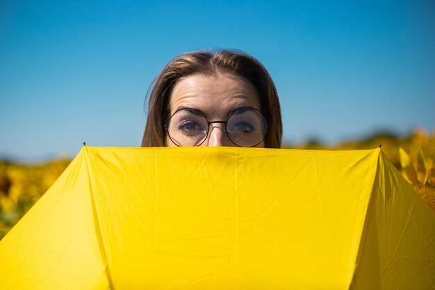 Jeune Femme Avec Des Lunettes Furtivement Derrière Un Parapluie Jaune Sur Un Champ De Tournesol.