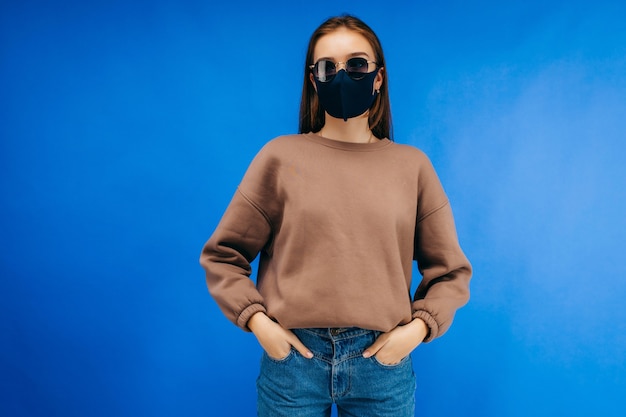 Jeune femme à lunettes, masque médical et sweat à capuche qui pose en studio sur fond bleu