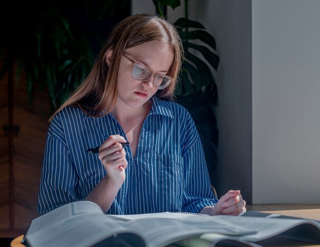 Jeune femme à lunettes pensant à la lecture se préparant aux examens avec des cahiers des livres au bureau en bois i...