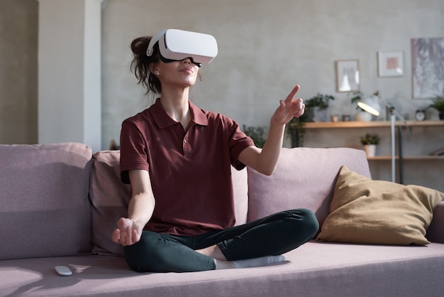 Jeune femme à lunettes de réalité virtuelle assise sur un canapé et jouant à un jeu vidéo à la maison