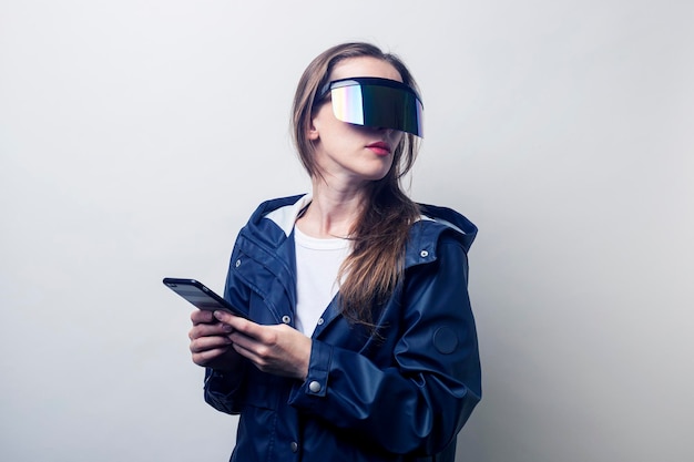 Jeune femme à lunettes de réalité virtuelle avec un téléphone dans une veste bleue sur fond clair