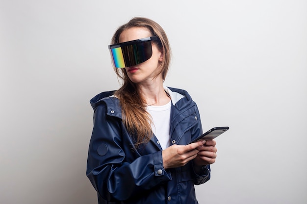 Jeune femme à lunettes de réalité virtuelle avec un téléphone dans une veste bleue sur fond clair.