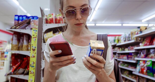 Jeune Femme à Lunettes De Soleil Debout Dans L'épicerie Et à L'aide De Téléphone En Vérifiant La Case De Boisson Fraîche