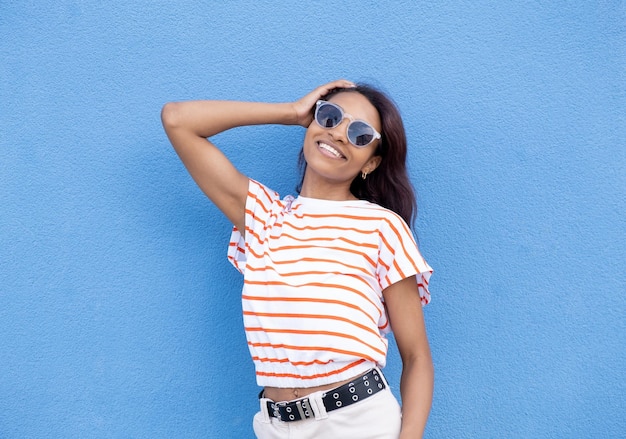 Jeune femme à lunettes de soleil élégantes avec des cheveux bouclés attachés en queue de cheval haute regardant loin tout en souriant largement montrant des dents droites parfaites posant sur fond de mur bleu