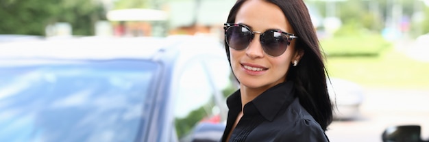 Jeune femme à lunettes de soleil se tient près d'une voiture noire dans un parking. Faites un essai routier avant d'acheter une voiture. Formation à la conduite