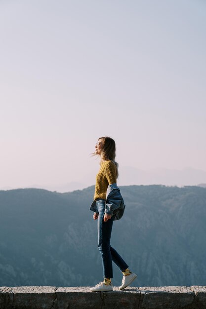 Une jeune femme en lunettes de soleil se tient sur un trottoir en pierre haut dans les montagnes vue latérale