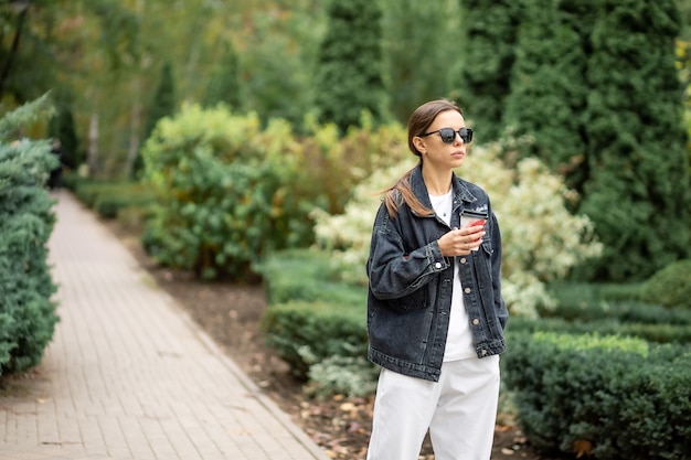 Jeune femme à lunettes de soleil et avec une tasse de café en papier dans le parc