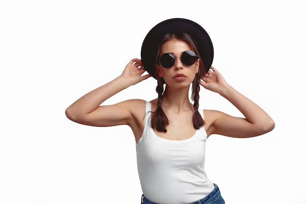 Jeune femme avec des lunettes de soleil tenant un chapeau avec les deux mains isolées sur un mur blanc