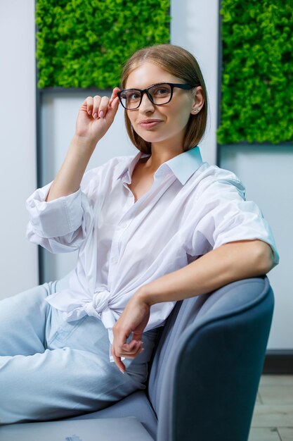 Une jeune femme à lunettes avec un sourire sur son visage, elle est dans une chemise blanche et un jean assis sur le canapé