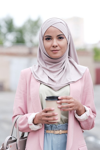 Jeune femme magnifique en hijab, pull décontracté, pantalon et cardigan rose prenant un café en se tenant debout dans un environnement urbain